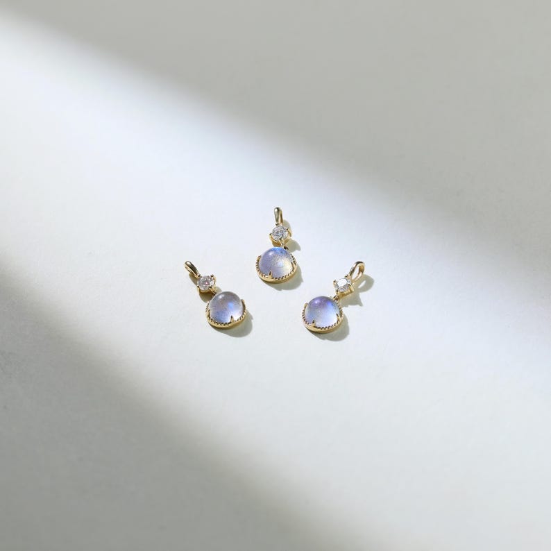 May include: Three gold pendants with a round, iridescent gemstone and a smaller clear gemstone. The pendants are on a white background.