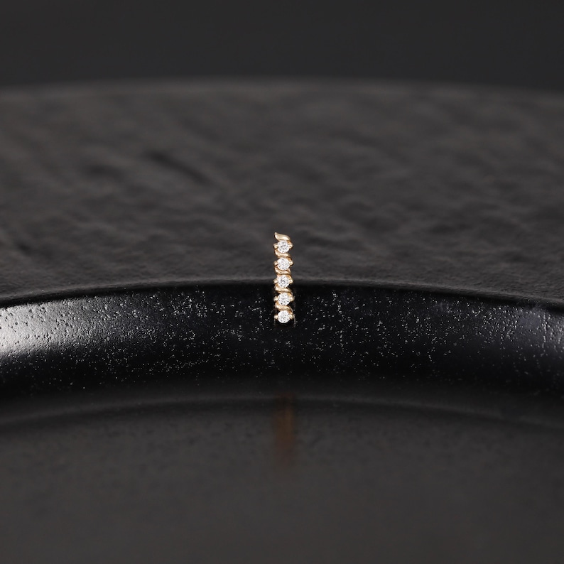 K&ouml;nnte beinhalten: Ein vertikaler Goldsteg-Ohrring mit f&uuml;nf kleinen, runden, klaren Steinen in einer Reihe. Das Schmuckst&uuml;ck hat eine polierte metallische Oberfl&auml;che und ein schlankes, l&auml;ngliches Design, das die Steine sicher entlang der Mittelachse h&auml;lt.