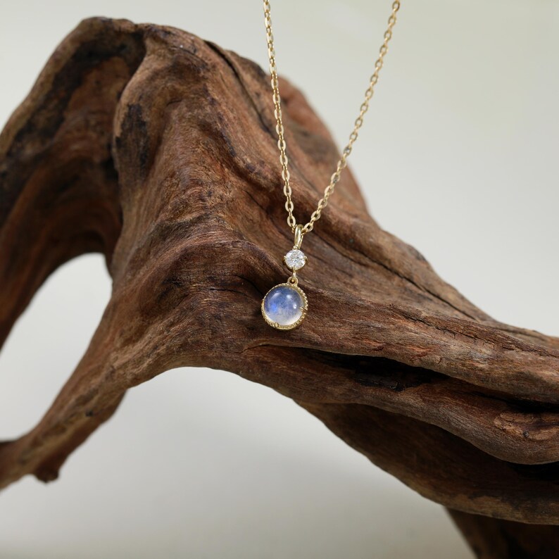 May include: A gold necklace with a round moonstone pendant and a smaller round diamond accent. The necklace is displayed on a piece of driftwood.