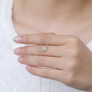 May include: A delicate gold necklace featuring a sunburst-shaped pendant with a central round iridescent stone. The pendant is suspended from a thin gold chain, showcasing a radiant design with small metallic rays extending from the center stone.