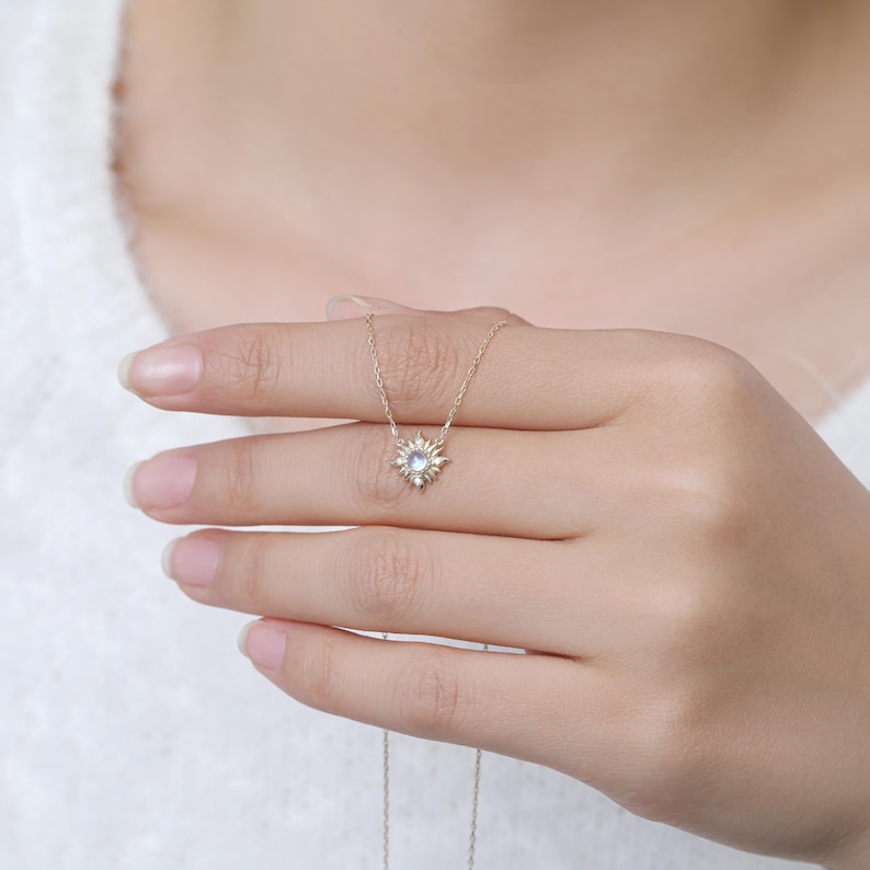 May include: A delicate gold necklace featuring a sunburst-shaped pendant with a central round iridescent stone. The pendant is suspended from a thin gold chain, showcasing a radiant design with small metallic rays extending from the center stone.