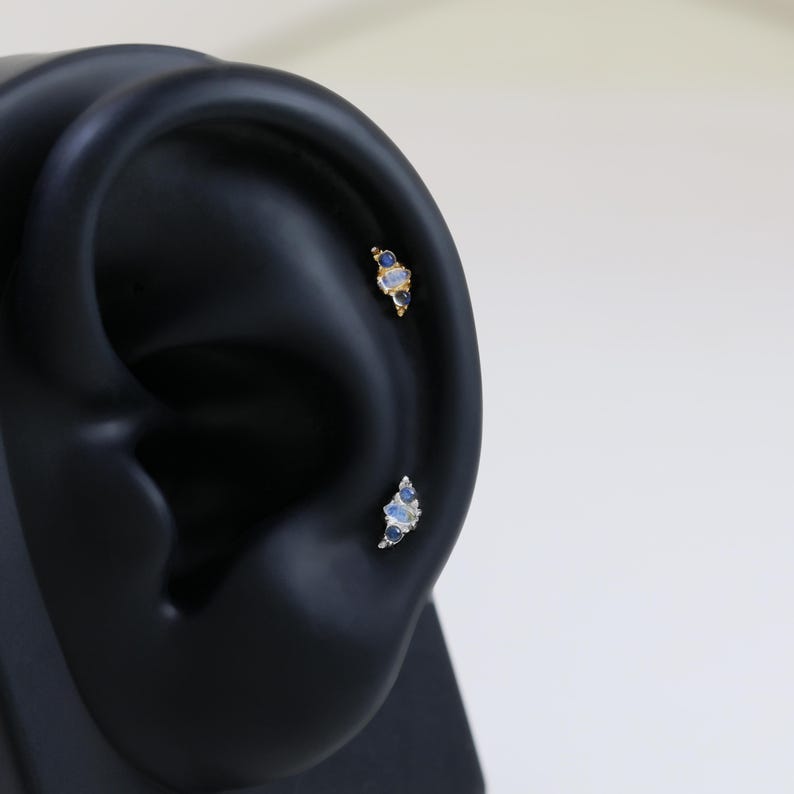 May include: Two gold-colored ear piercings with blue and white gemstones. The top piercing has a cluster of three stones, while the bottom one has a crescent-shaped arrangement of stones. The piercings are displayed on a black ear model.