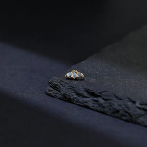 May include: A gold-colored jewelry piece featuring three light blue gemstones. The gemstones are set in a cluster design, with a small, textured gold accent between each stone. The piece is displayed on a dark, textured surface.