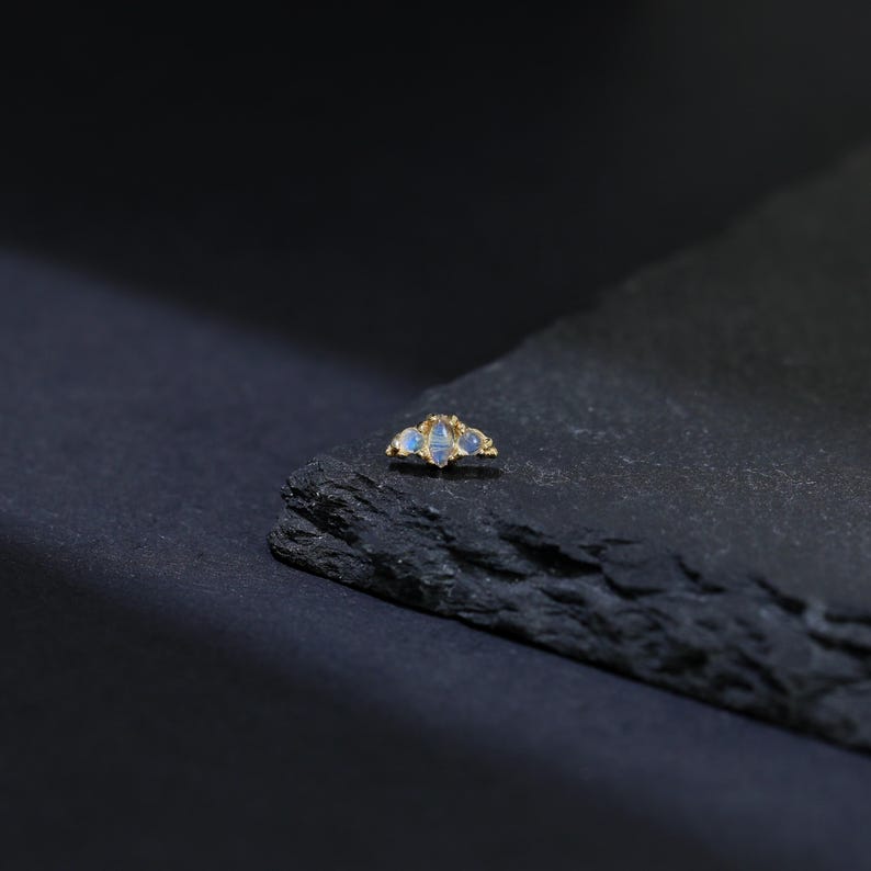May include: A gold-colored jewelry piece featuring three light blue gemstones. The gemstones are set in a cluster design, with a small, textured gold accent between each stone. The piece is displayed on a dark, textured surface.