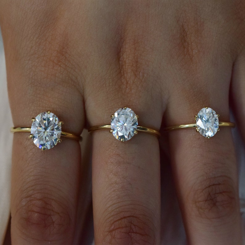 May include: Three gold rings with round, clear gemstones. The rings are on a person's fingers.