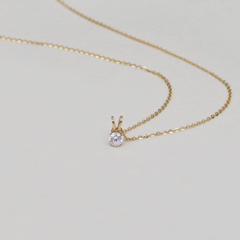 Pu&ograve; includere: Una delicata collana d'oro con un piccolo pendente rotondo di pietra preziosa trasparente. Il pendente &egrave; tenuto da un'incastonatura d'oro e pende da una sottile catena d'oro. La collana &egrave; esposta su uno sfondo morbido e neutro.