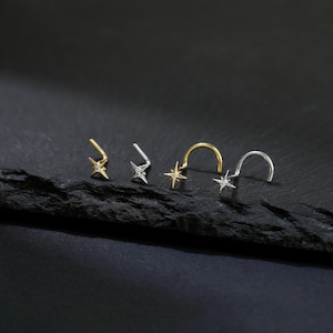 May include: Four nose rings with star-shaped charms. Two are gold-toned, and two are silver-toned. The star charms have small, clear stones. The nose rings are displayed on a dark, textured surface against a black background.