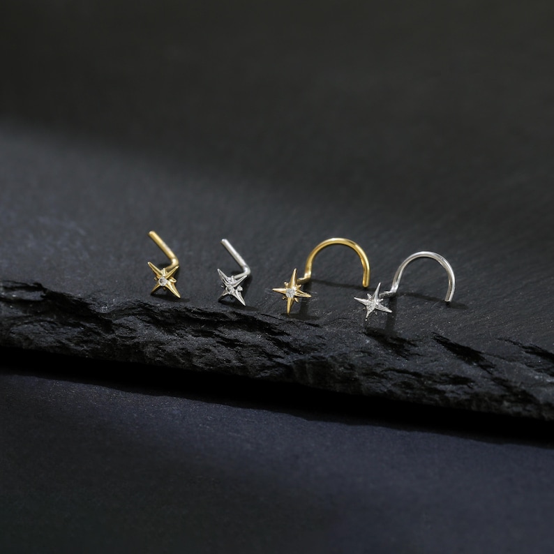 May include: Four nose rings with star-shaped charms. Two are gold-toned, and two are silver-toned. The star charms have small, clear stones. The nose rings are displayed on a dark, textured surface against a black background.