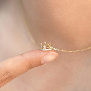 May include: A gold necklace featuring a small crown-shaped pendant. The crown is embellished with tiny, clear, sparkling stones along its base and points, attached to a delicate gold chain that rests against the skin.