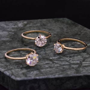 May include: Three gold engagement rings with round, clear diamonds on a dark green marble surface.