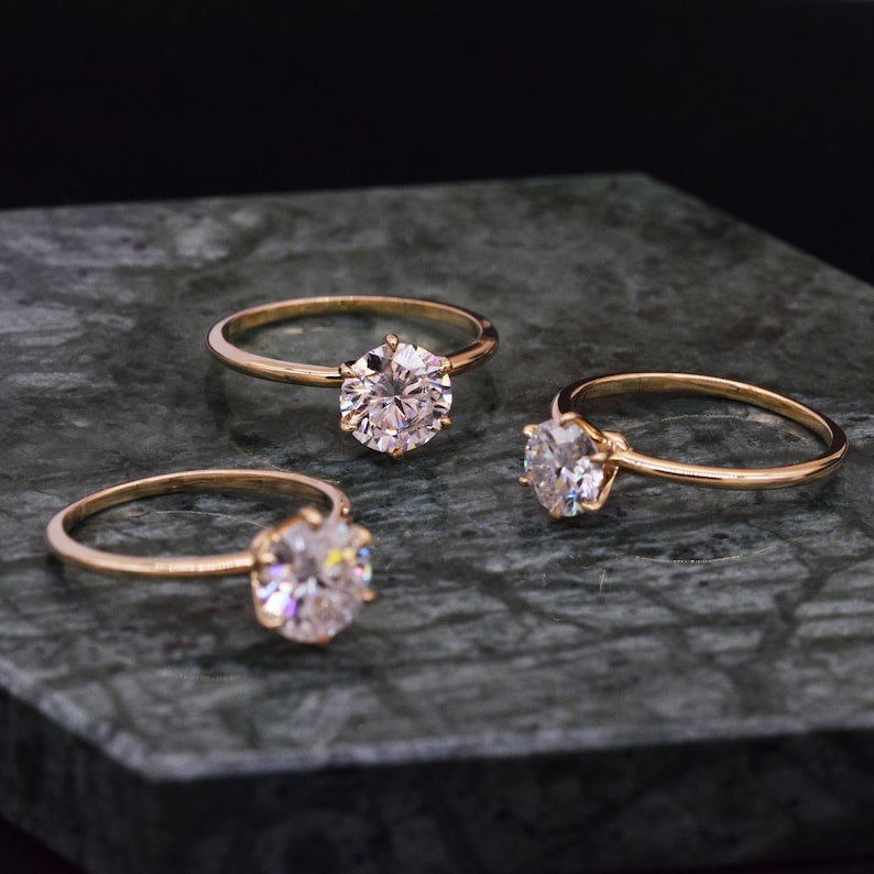 May include: Three gold engagement rings with round, clear diamonds on a dark green marble surface.