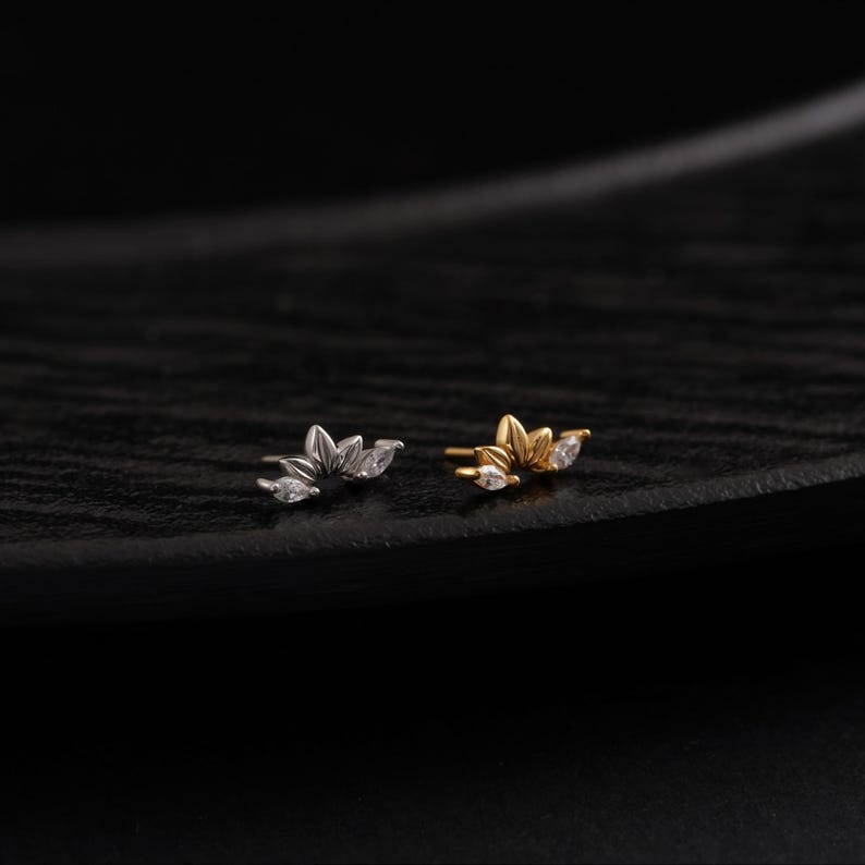 May include: Two small, decorative earrings. One is silver-toned, the other gold-toned. Each earring features a floral design with three clear, marquise-cut gemstones. The earrings are set against a dark, textured background.