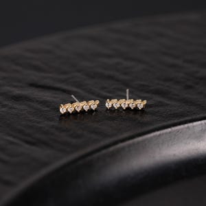 K&ouml;nnte beinhalten: Ein Paar goldene Ohrstecker, jeweils mit einem horizontalen Balkendesign, das mit f&uuml;nf kleinen, klaren, rund geschliffenen Steinen besetzt ist. Die Ohrringe haben ein metallisch goldenes Finish und einen d&uuml;nnen Metallstift an der R&uuml;ckseite.