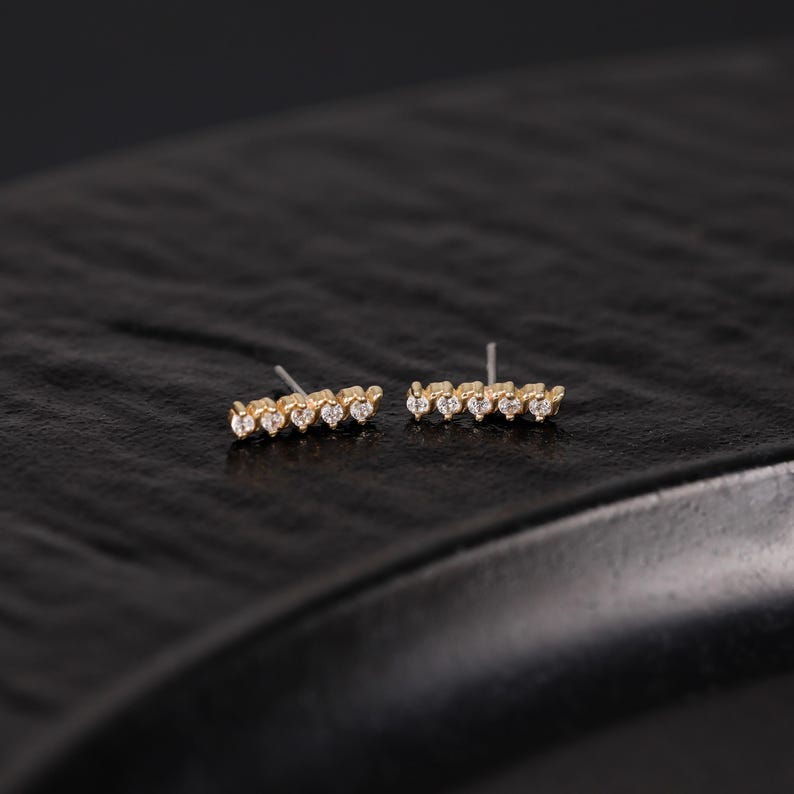 K&ouml;nnte beinhalten: Ein Paar goldene Ohrstecker, jeweils mit einem horizontalen Balkendesign, das mit f&uuml;nf kleinen, klaren, rund geschliffenen Steinen besetzt ist. Die Ohrringe haben ein metallisch goldenes Finish und einen d&uuml;nnen Metallstift an der R&uuml;ckseite.