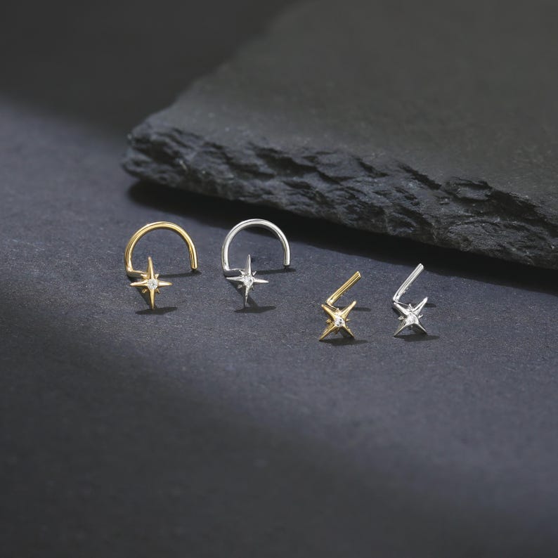 May include: Four pieces of jewelry: two nose rings and two ear studs. The nose rings are gold and silver, each with a star-shaped charm and a small clear gem. The ear studs are gold and silver, each with a star-shaped charm and a small clear gem.