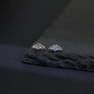 May include: Two pieces of jewelry, each with three light blue, teardrop-shaped stones. One piece has a gold-colored setting, and the other has a silver-colored setting. The jewelry is displayed on a dark, textured surface.