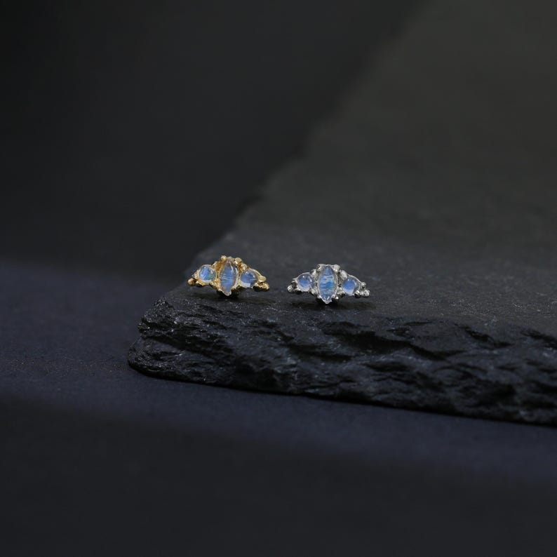 May include: Two pieces of jewelry, each with three light blue, teardrop-shaped stones. One piece has a gold-colored setting, and the other has a silver-colored setting. The jewelry is displayed on a dark, textured surface.