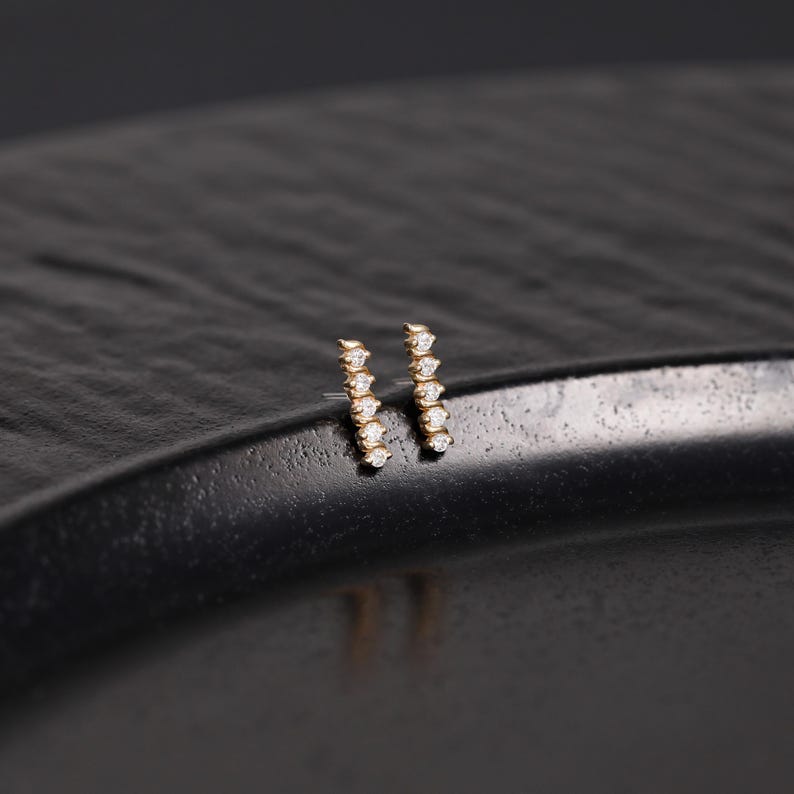 K&ouml;nnte beinhalten: Ein Paar Ohrstecker aus Gold, jeweils mit einem vertikalen Stabdesign, das mit f&uuml;nf kleinen, klaren, rund geschliffenen Steinen in einer Reihe besetzt ist. Die Ohrringe haben eine polierte metallische Oberfl&auml;che und eine schlichte Form.