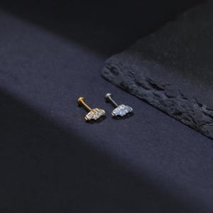 May include: Two small stud earrings. One is gold-toned, the other is silver-toned. Each earring features three small, light-colored gemstones arranged in a curved design. The earrings are displayed on a dark surface.