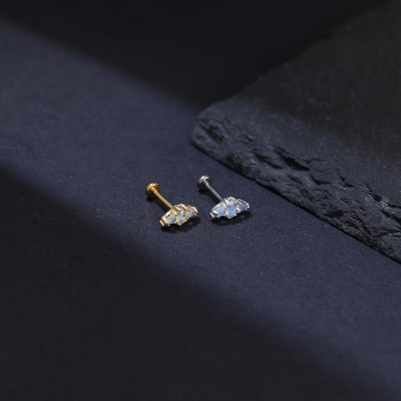May include: Two small stud earrings. One is gold-toned, the other is silver-toned. Each earring features three small, light-colored gemstones arranged in a curved design. The earrings are displayed on a dark surface.
