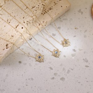 May include: Three gold necklaces featuring sunburst-shaped pendants. Each pendant has a central round iridescent stone surrounded by small clear stones. The delicate gold chains are thin and link-style, extending from the sun-shaped charms.