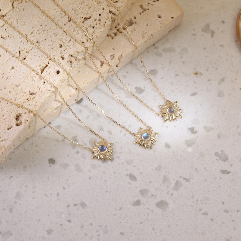 May include: Three gold necklaces featuring sunburst-shaped pendants. Each pendant has a central round iridescent stone surrounded by small clear stones. The delicate gold chains are thin and link-style, extending from the sun-shaped charms.
