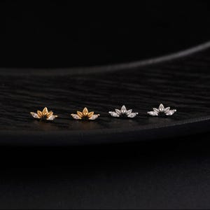 May include: Four pairs of small, gold and silver-toned lotus flower stud earrings with clear, marquise-cut cubic zirconia accents. The earrings are displayed on a dark, textured surface, showcasing their delicate design and sparkle.