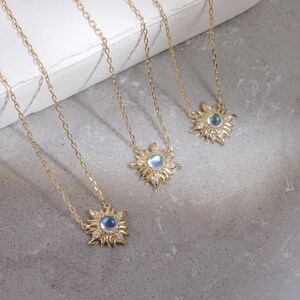 May include: Three gold necklaces featuring sunburst-shaped pendants. Each pendant has a central round iridescent moonstone cabochon surrounded by small clear stones set in a radiating metal frame. The necklaces have delicate gold link chains.