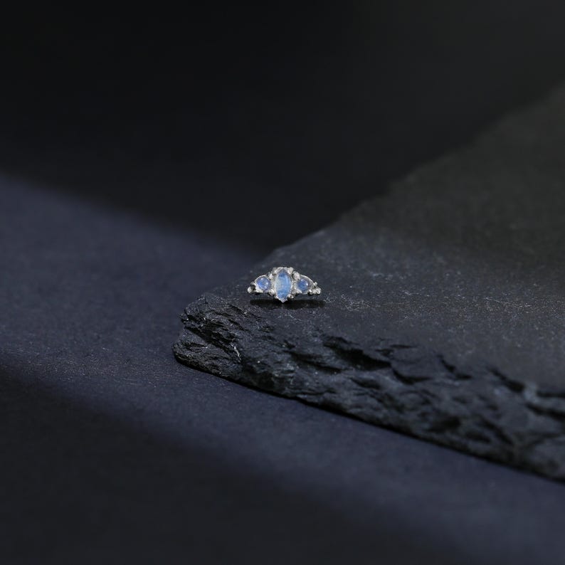 May include: A silver ring with three light blue oval-shaped gemstones. The ring is set on a dark gray, textured surface against a dark background. The gemstones are the focal point of the ring.
