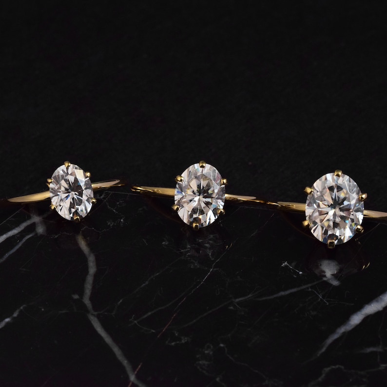 May include: Three gold rings with clear round gemstones on a black marble surface.