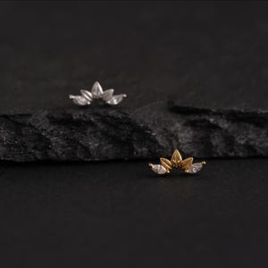 May include: Two small, decorative ear studs. One is silver-toned, the other is gold-toned. Each stud features a curved design with three marquise-cut, clear gemstones. The studs are displayed on a dark, textured surface.