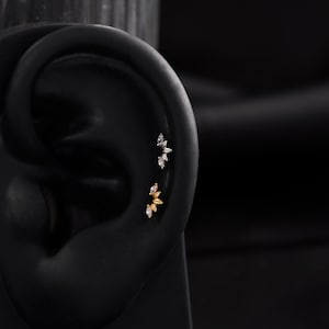 May include: Close-up of an ear with two sets of decorative ear piercings. The top set is silver-toned, and the bottom set is gold-toned. Each set features a floral design with small, clear gemstones. The ear is black, and the background is dark.
