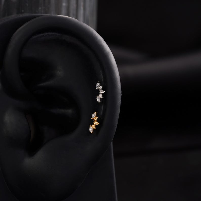 May include: Close-up of an ear with two sets of decorative ear piercings. The top set is silver-toned, and the bottom set is gold-toned. Each set features a floral design with small, clear gemstones. The ear is black, and the background is dark.