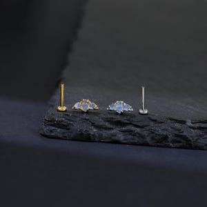 May include: A collection of three pieces of jewelry, including two lip or ear studs and a pair of earrings. The earrings and studs feature a gold or silver setting with three light blue, teardrop-shaped stones. The jewelry is displayed on a dark, textured surface.
