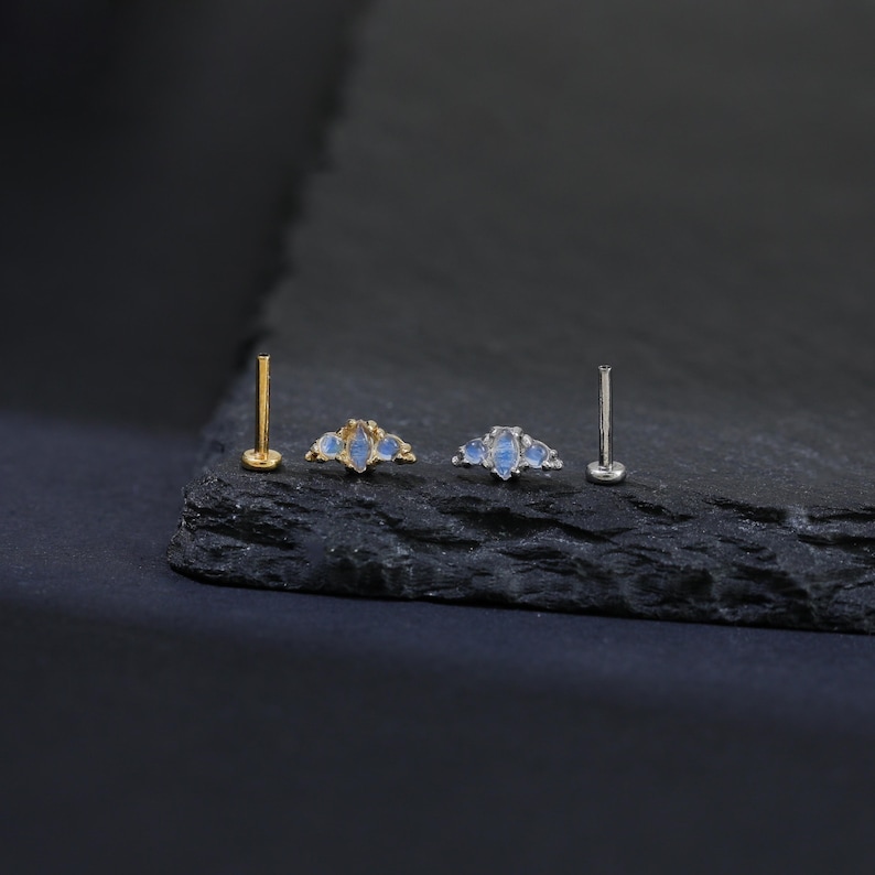 May include: A collection of three pieces of jewelry, including two lip or ear studs and a pair of earrings. The earrings and studs feature a gold or silver setting with three light blue, teardrop-shaped stones. The jewelry is displayed on a dark, textured surface.