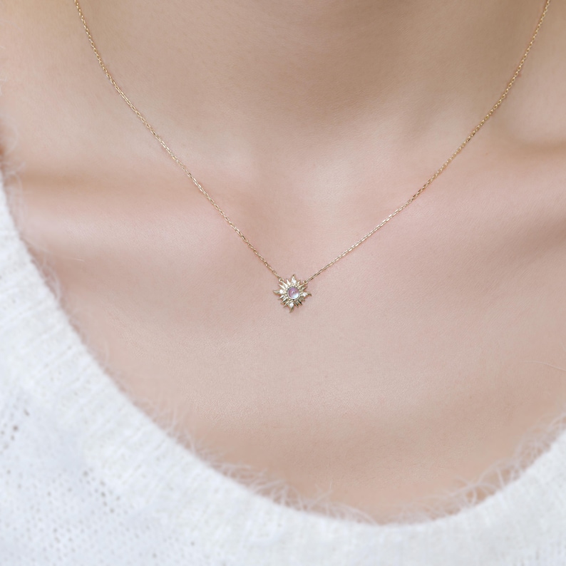 May include: A delicate gold chain necklace featuring a small, starburst-shaped pendant. The center of the pendant contains a single, round, light purple gemstone surrounded by tiny, clear, sparkling stones set in a sunray-inspired metal frame.