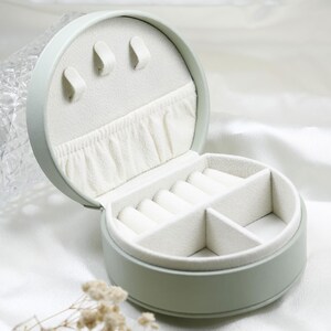May include: A round, sage green jewelry box with a cream-colored interior. The lid features three hooks and a gathered pocket, while the base contains a ring roll section and two open compartments for organizing small accessories and jewelry items.