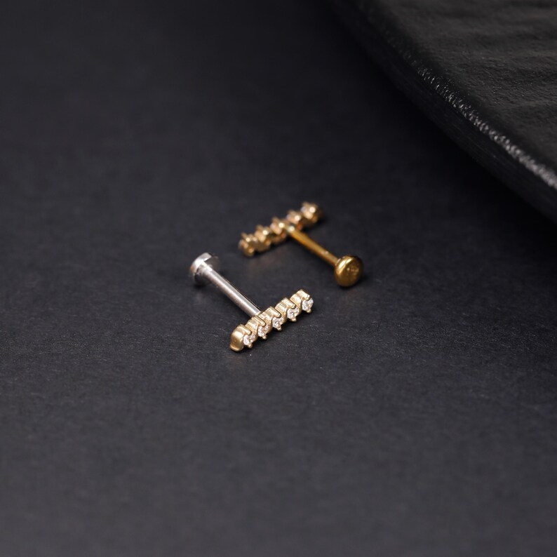 K&ouml;nnte beinhalten: Zwei Labret-Piercings mit flacher R&uuml;ckseite, eines in Silberoptik und eines in Goldoptik. Jedes Piercing besteht aus einem horizontalen Stab mit f&uuml;nf kleinen, klaren, runden Steinen in einer Reihe, befestigt an einem d&uuml;nnen Stift mit flacher Scheibe.