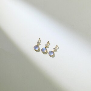 May include: Three gold pendant charms with a round, iridescent blue gemstone and a small, clear crystal accent.