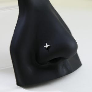 May include: A silver-colored nose stud in the shape of a four-pointed star. The stud is set on a black mannequin nose, with a white background. The star has a shiny, reflective surface.
