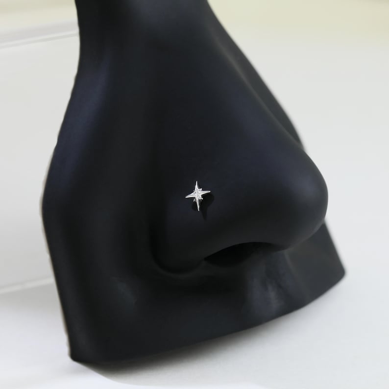 May include: A silver-colored nose stud in the shape of a four-pointed star. The stud is set on a black mannequin nose, with a white background. The star has a shiny, reflective surface.