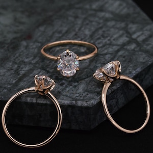May include: Three gold engagement rings with oval-shaped diamonds. The rings are on a dark gray textured surface.