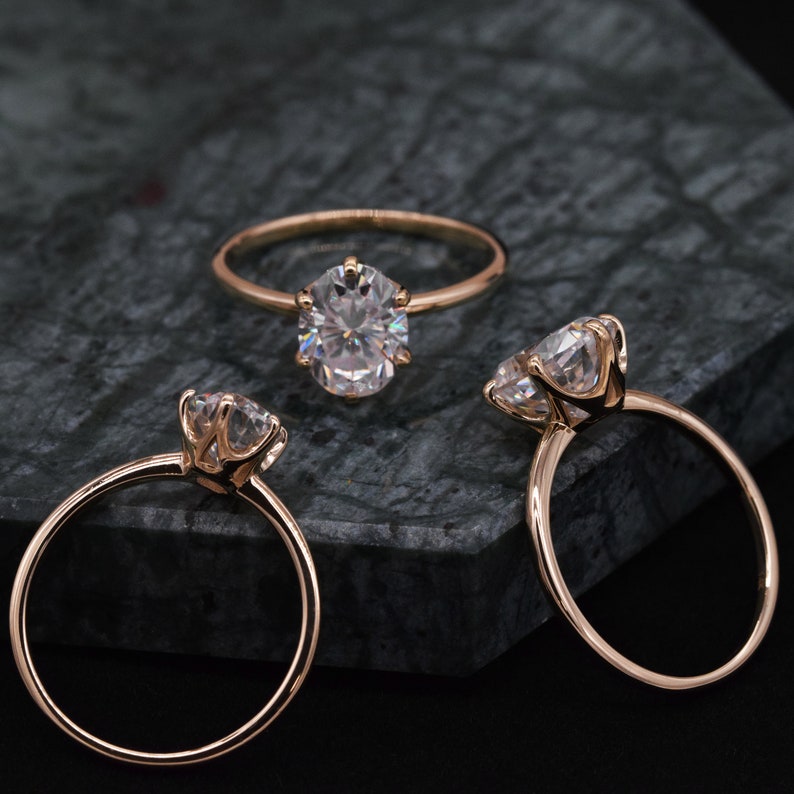 May include: Three gold engagement rings with oval-shaped diamonds. The rings are on a dark gray textured surface.