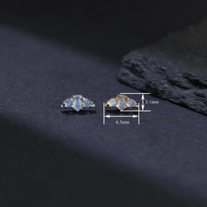 May include: Two small, gold-toned earrings with a crescent shape, each adorned with three oval, light blue gemstones. The earrings are 6.5 mm wide and 3.1 mm high. The background is dark.