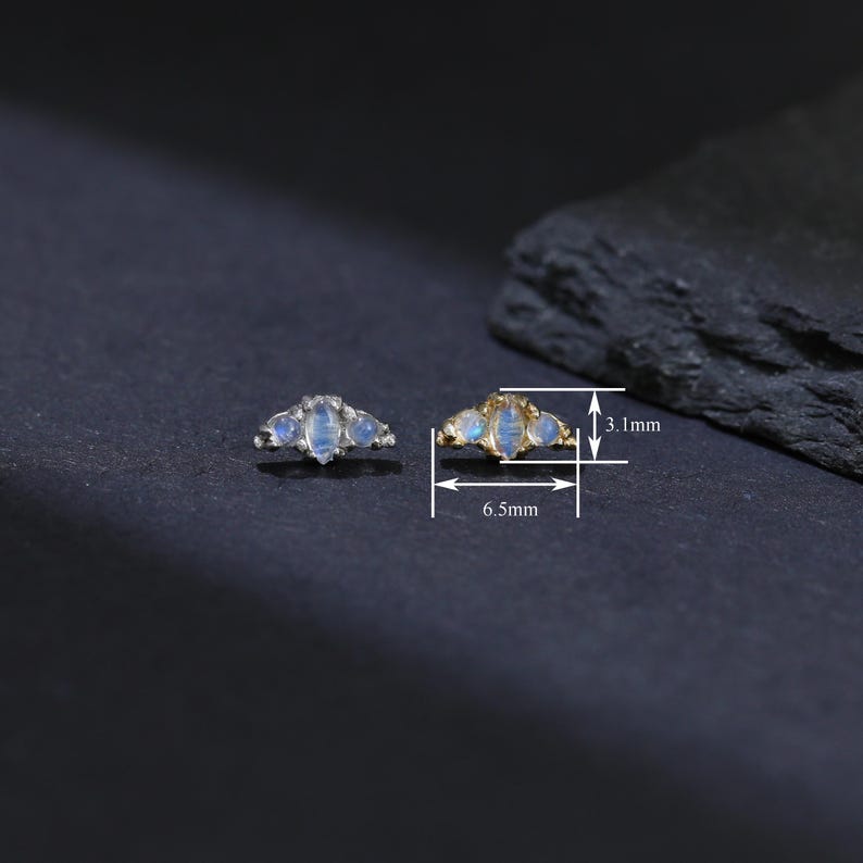 May include: Two small, gold-toned earrings with a crescent shape, each adorned with three oval, light blue gemstones. The earrings are 6.5 mm wide and 3.1 mm high. The background is dark.