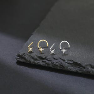 May include: Four star-shaped nose rings, two gold and two silver, each with a small, clear gemstone at the center. The nose rings are displayed on a dark, textured surface, showcasing their delicate design and sparkle.