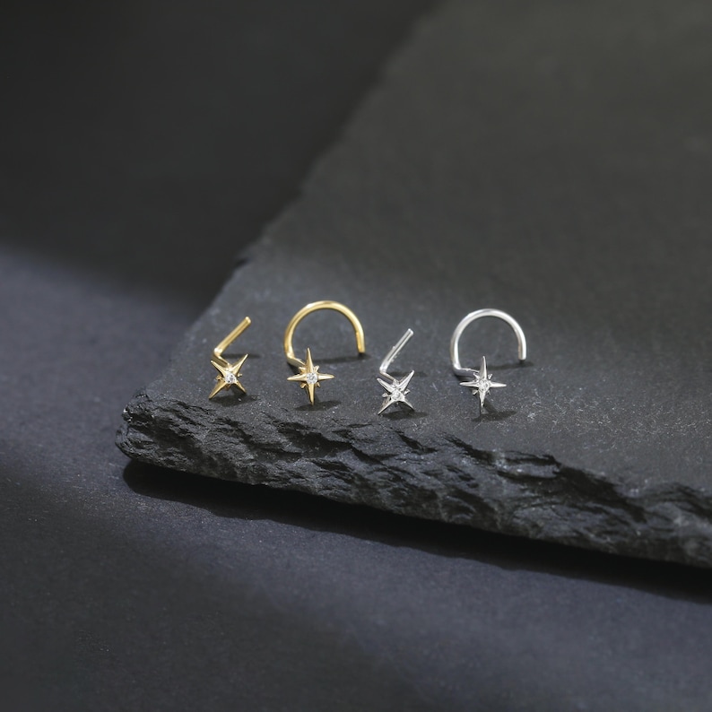 May include: Four star-shaped nose rings, two gold and two silver, each with a small, clear gemstone at the center. The nose rings are displayed on a dark, textured surface, showcasing their delicate design and sparkle.