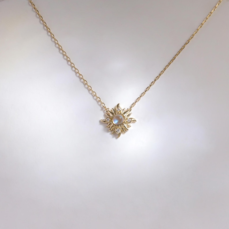 May include: A gold chain necklace featuring a sun-shaped pendant. The pendant has a central round iridescent stone surrounded by radiating gold rays and small clear stones set within the metal points of the sun design.