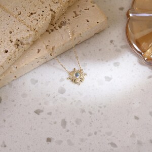 May include: A gold-toned necklace featuring a delicate chain and a sunburst-shaped pendant. The center of the pendant holds a small, iridescent round stone, surrounded by radiating metal rays that create a star-like appearance on the jewelry piece.