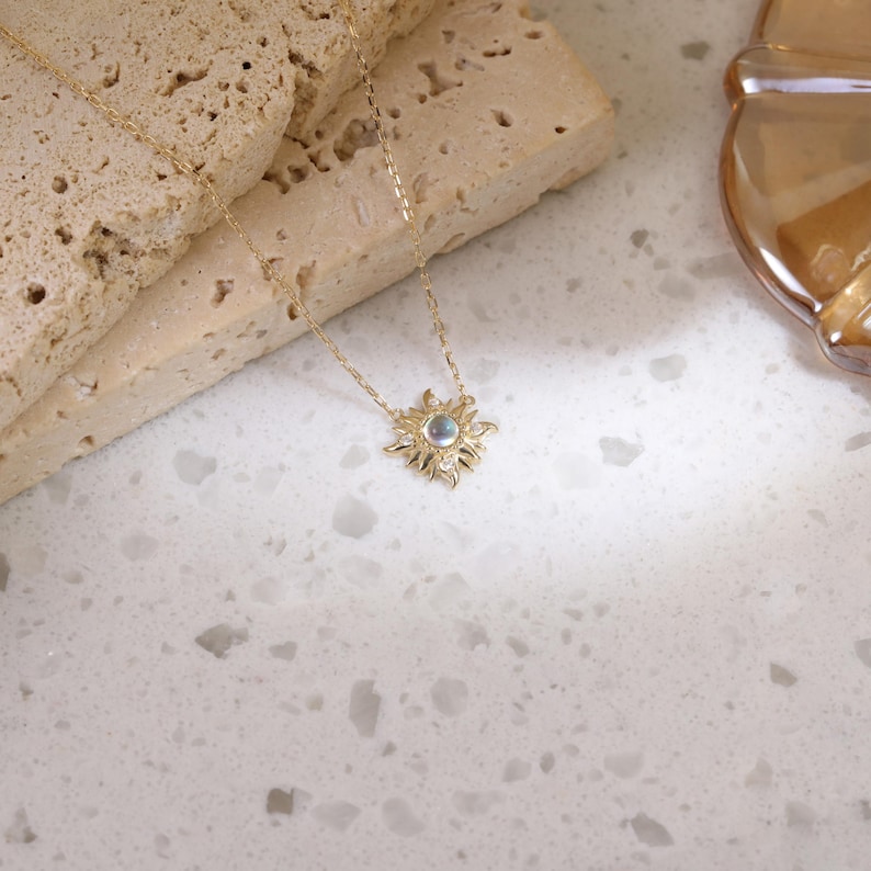 May include: A gold-toned necklace featuring a delicate chain and a sunburst-shaped pendant. The center of the pendant holds a small, iridescent round stone, surrounded by radiating metal rays that create a star-like appearance on the jewelry piece.